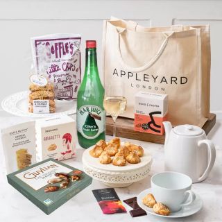 A selection of sweet and savoury foods places around a bottle, white teapot and cup and saucer, and a large Appleyard London material bag