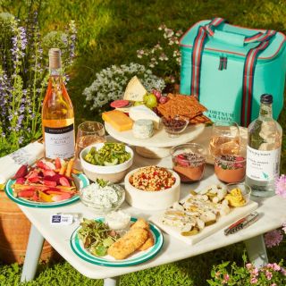 A selection of sweet and savoury food on plates, bowls and cheeseboards, with a bottle of rosé wine on a table in front of teal Fortnum and Mason picnic hamper. It is surrounded by flowers and shrubs