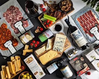 A bird's eye view of a selection of charcuterie foods include cured meats, cheeses, a tin of olives, salad and a bottle of wine