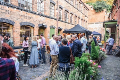 A courtyard of a factory with shrubbery and several gazebos with lots of people standing around in groups