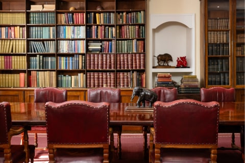 Red leather embossed chairs surround a wooden table against a wooden bookcase filled with old books. There is a large elephant ornament on the table