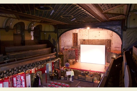 The view from the back of a hall with pew seating upstairs and seats and panels on the ground with the stage at one end