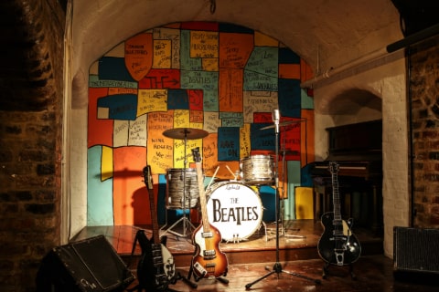 The inside of a room with a drum kit with The Beatles logo on it, several guitars and a mike stand with coloured patches on the wall