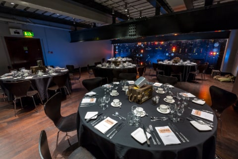 A room with several tables set for an event with 8 settings per table and a stage in the background