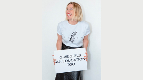 A colour portrait of Juliet Kinsman looking away from the camera while holding a sign which reads give girls and education too. Juliet is a white woman with blonde hair wearing a white t-shirt and black skirt.