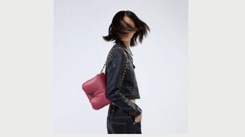  A white model swings her hair covering her face, She wears a denin top and jeans and has a pink cushioned bag over her shoulder 