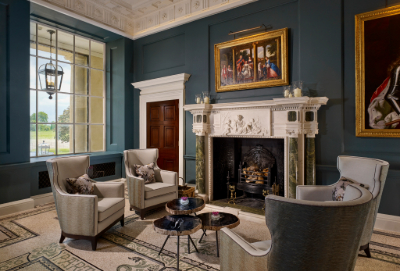 A grey painted room with a white ornate fireplace.  There are oil paintings on the wall, a large glass window, a brown door, 4 pale armchairs facing each other around three small coffee tables.