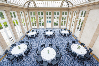 A glass ceilinged room with glass doors holds 6 round tables with 8 chairs at each one.