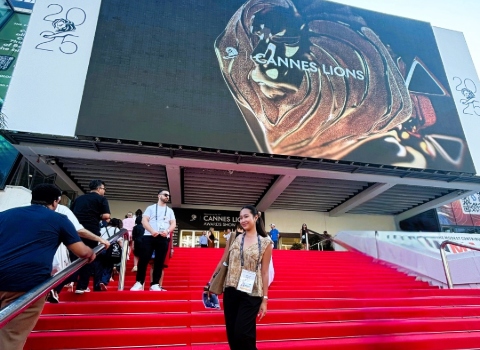 Leticia Callista stood on red carpeted stairs which lead to a conference venue. Above her on the side of the venue is an advert for Cannes Lions 2025.