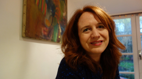 A colour photograph of Kate Hinton. Kate is a white woman with light brown shoulder length hair. She is sat in front of a picture and glass doors.