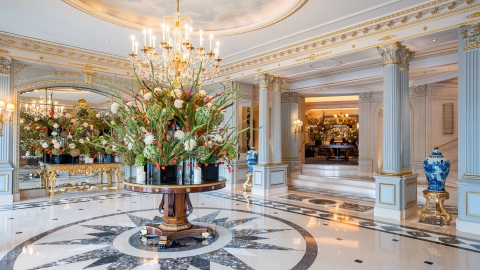 The ornate lobby features gold-etched columns and a floral centrepiece under a large ceiling-hung chandelier.