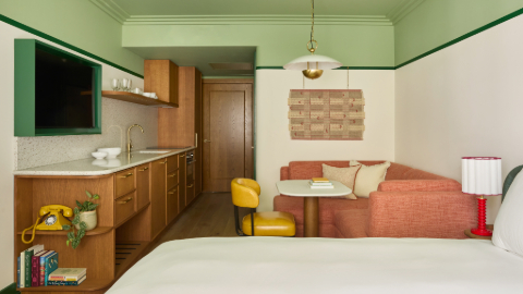 A bedroom with pistachio ceiling, white walls, bed, table, yellow chair and pink L-shaped sofa, wooden kitchen cupboards, white worktop and white bed.
