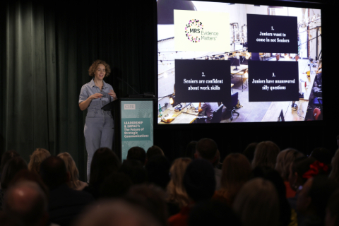 Naomi Goodman addressing the audience in the dark auditorium. To the right is a power point demonstration on a large screen.