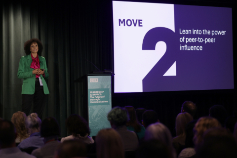 Naomi Goodman addressing the audience in the dark auditorium. To the right is a screen with a large number two and the words lean into the power of peer-to-peer influence