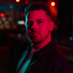 A colour portrait of Josh Wheeler under a dark red glow. Josh is a white man with short dark hair, wearing a dark jacket.