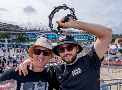 Two white males of different ages wearing white and black buckets hats and black Oasis-branded t-shirts outside a stadium