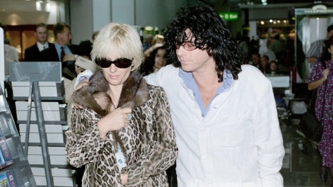 Paula Yates wearing a sunglasses and leopard print coat walking alongside Michael Hutchence who is wearing a white jacket and has his arm around her shoulder. 