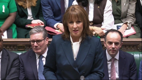 A colour photograph of Rachel Reeves stood in the House of Commons. Keir Starmer and other ministers and MPs are sat on the green benches behind her.
