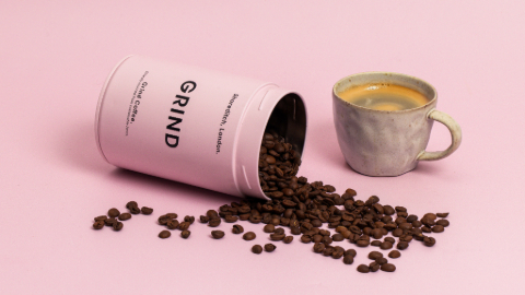 On a pink background a pink metallic jar with the word Grind on it, lies on its side. Coffee beans are scattered from cylinder to the pink surface. A cup of coffee is placed to the right.