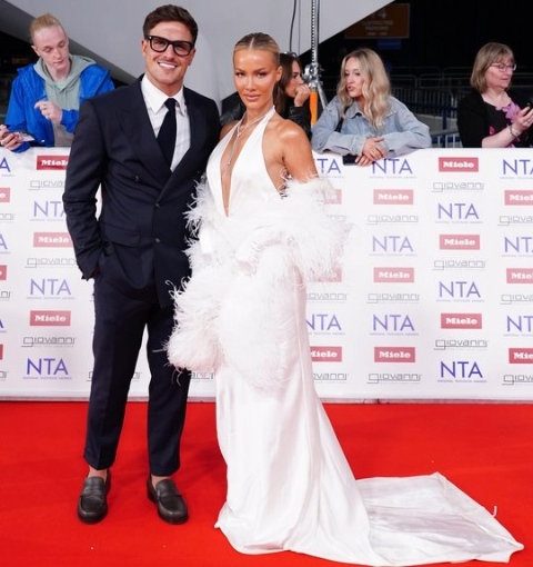 On a red carpet, Luca Bish wearing a black suit and tie smiles, while stood with Grace Jackson who is wearing a long white, sleeveless dress with train. In the background behind a barrier stands four fans.