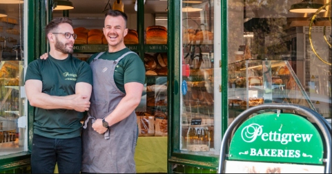Two smiling white men - both wearing green t-shirts, one partly covered by a grey apron - stand outside a bakery shop with green framed windows. A green sign with the company name is visible to the right.