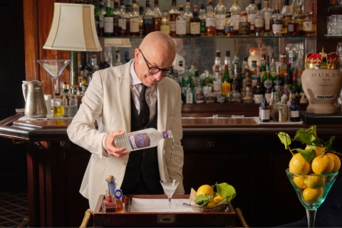 A male mixologist wearing a white jacket pours a martini in a bar.
