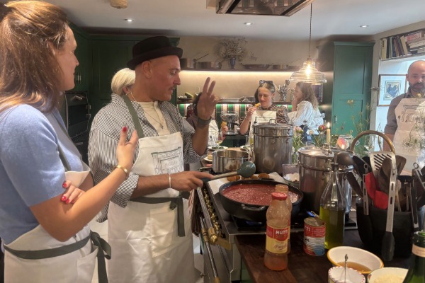 A white woman and white man stand in front of a bowl of pasta sauce on a hub in a large kitchen. Other people can be seen cooking in the background.