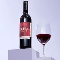 A bottle of red wine with red label on a plinth next to a glass of red wine.